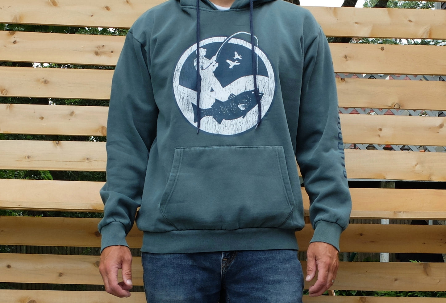 man wearing washed blue-green hoodie with round white and navy blue surf fisherman logo