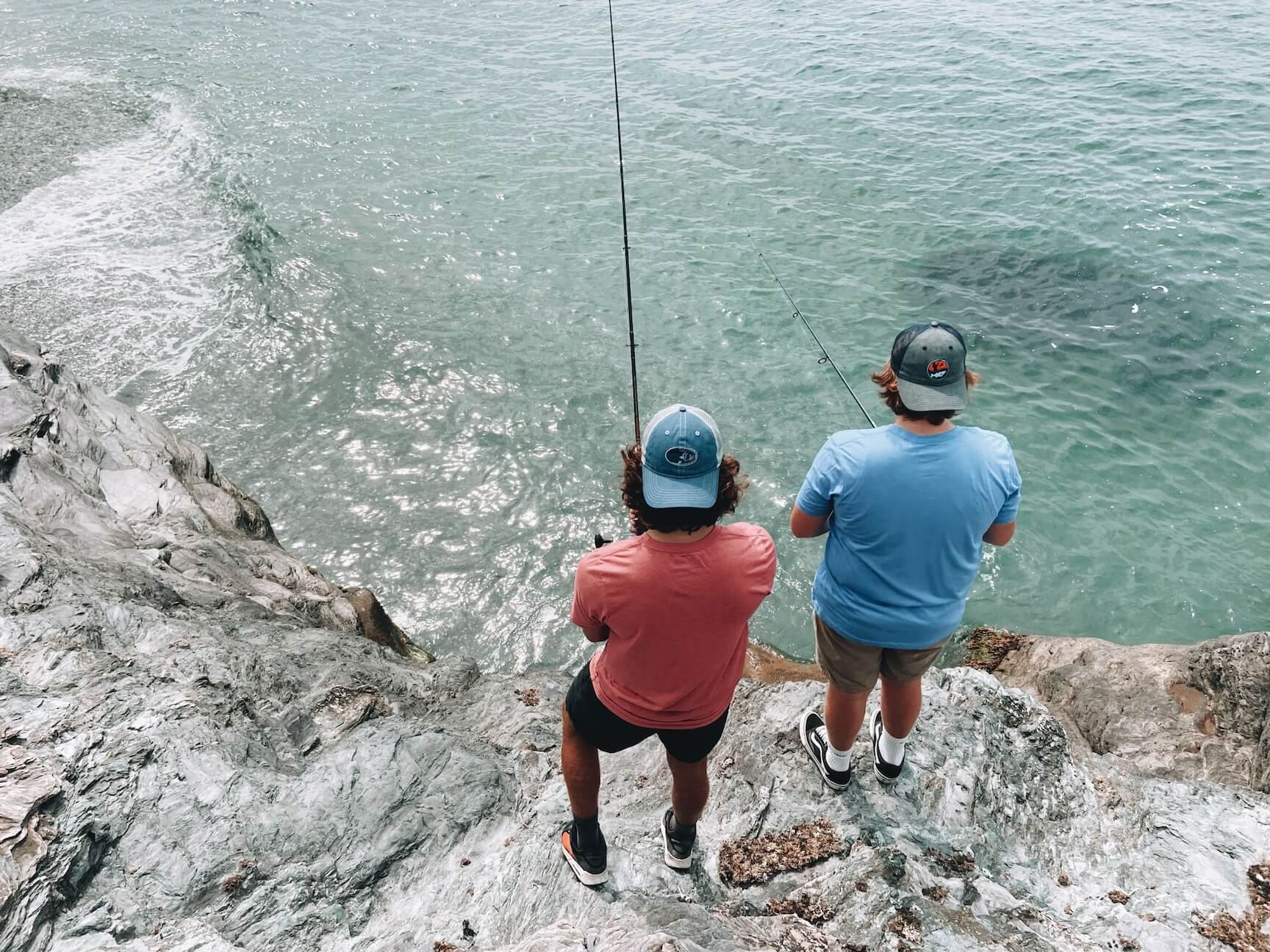 back view of two young men fishing from ocean rocks both wearing Deep Thoughts Designs fishing t-shirts