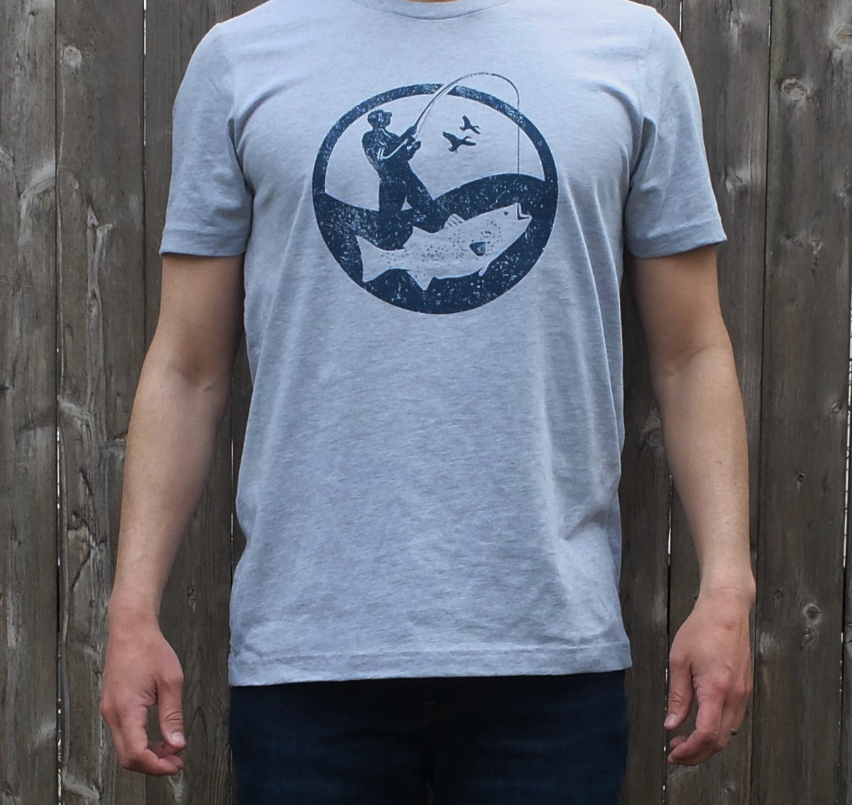 man wearing heather grey t-shirt with round navy blue surf fisherman logo graphic