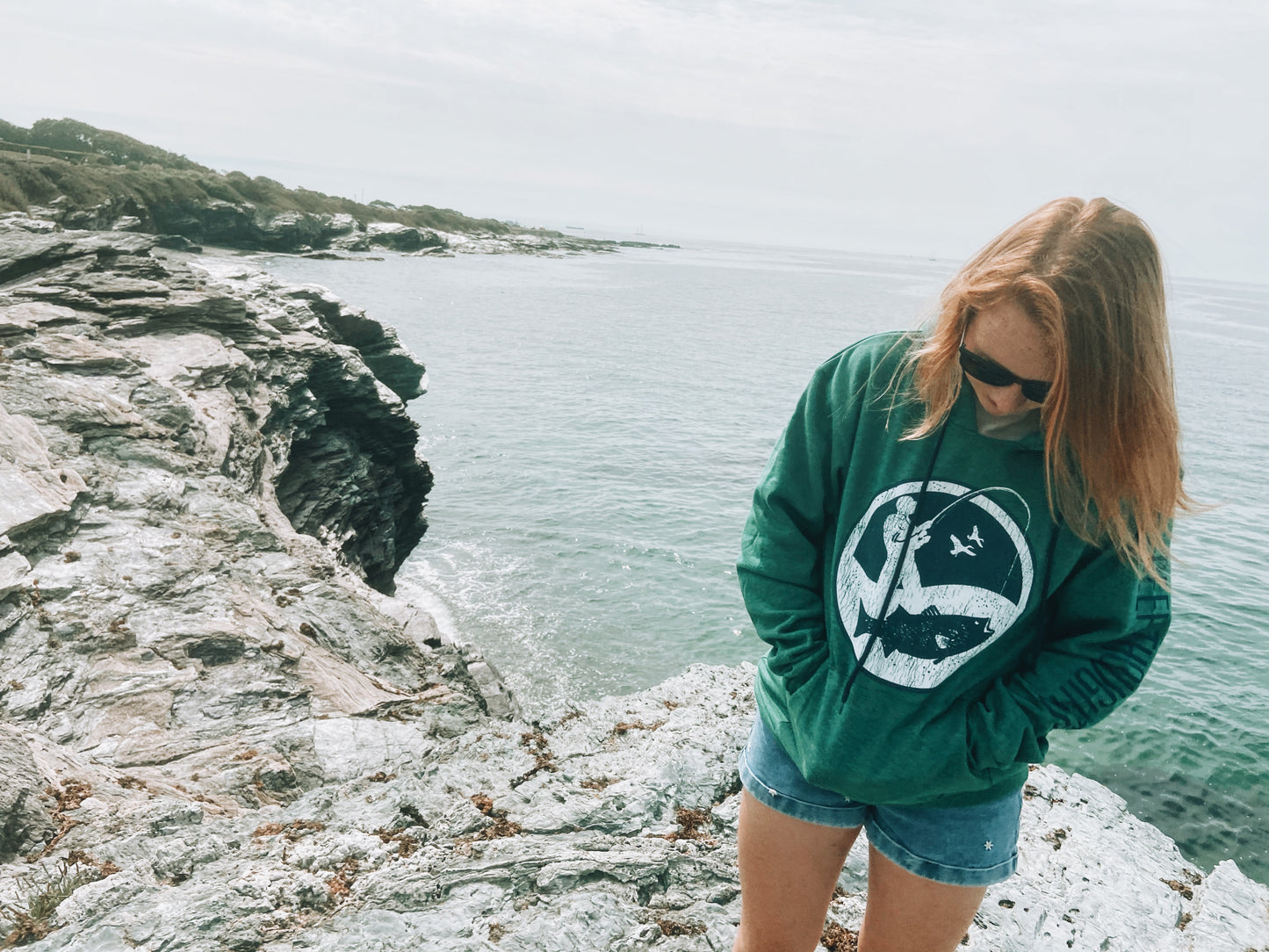 woman wearing heather green hoodie with large round navy blue and white surf fisherman logo