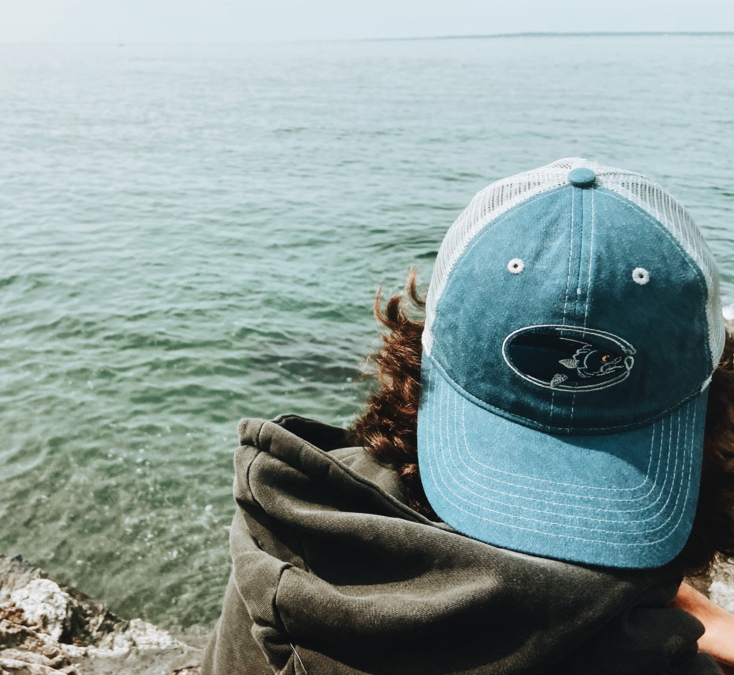 man wearing embroidered blue and grey bluefish trucker cap by the ocean
