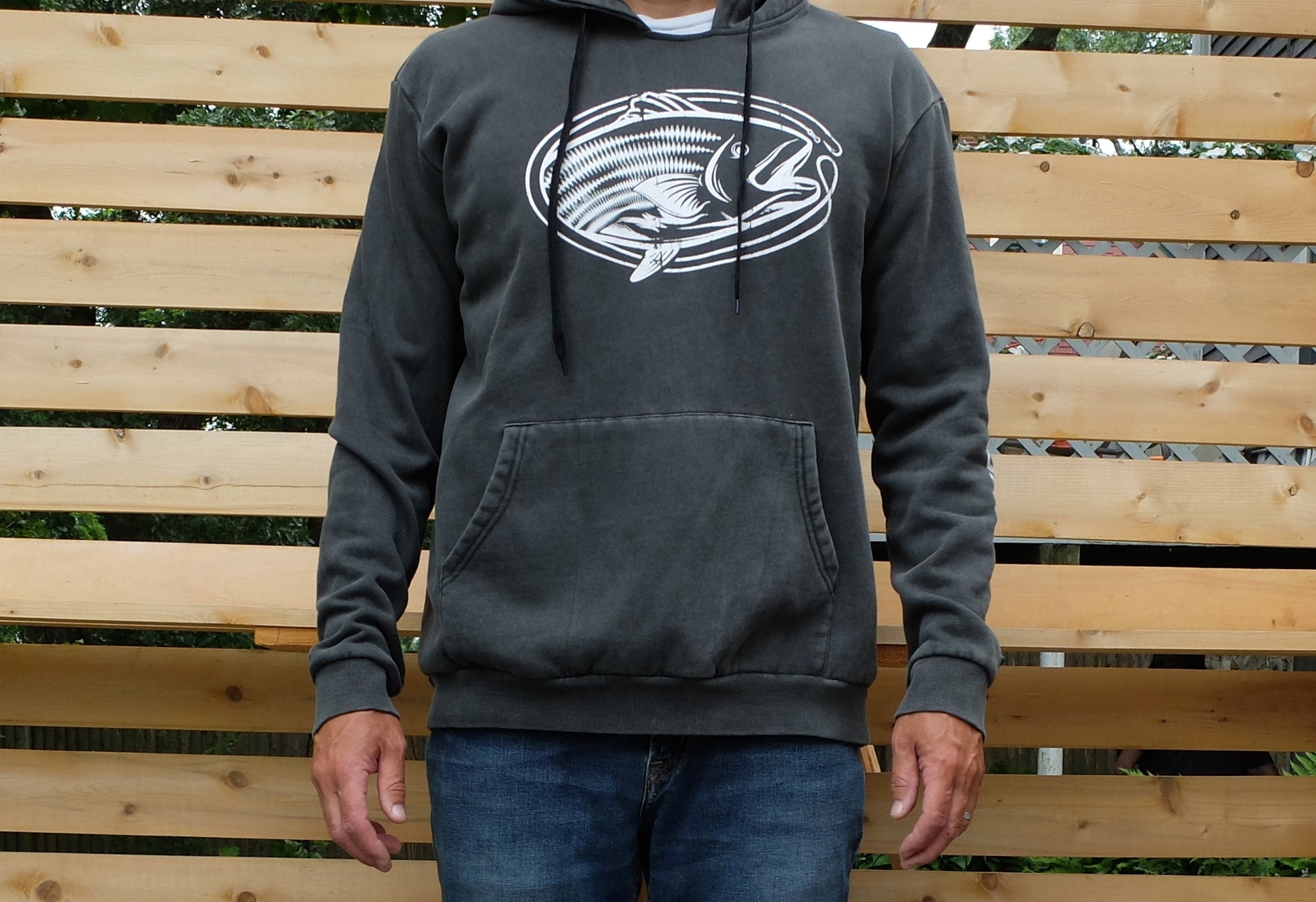 man wearing washed black garment dye hoodie with white vintage style oval-shaped striped bass graphic